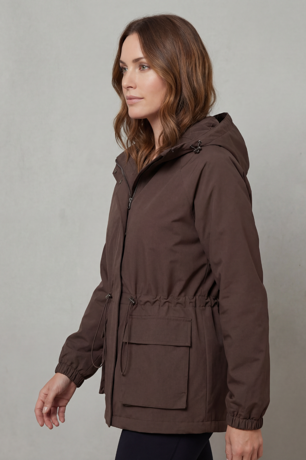 Modella indossa una giacca impermeabile marrone con cappuccio, coulisse in vita, tasche utility e chiusura con zip, su sfondo neutro_2