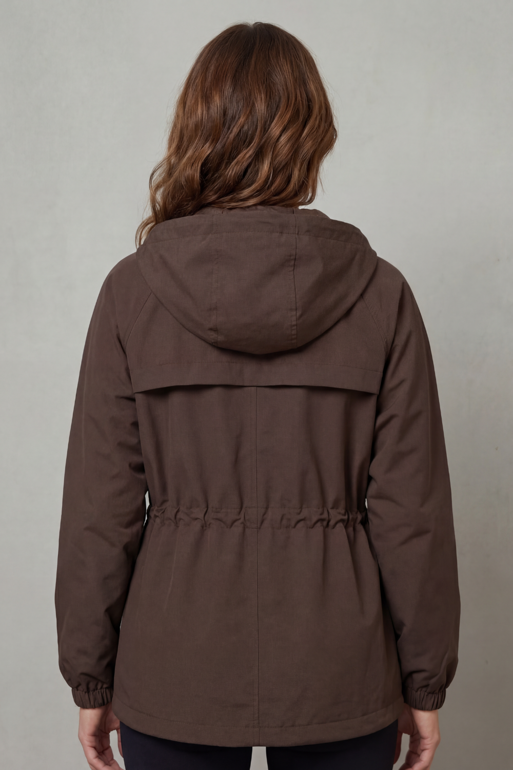 Modella indossa una giacca impermeabile marrone con cappuccio, coulisse in vita, tasche utility e chiusura con zip, su sfondo neutro_3